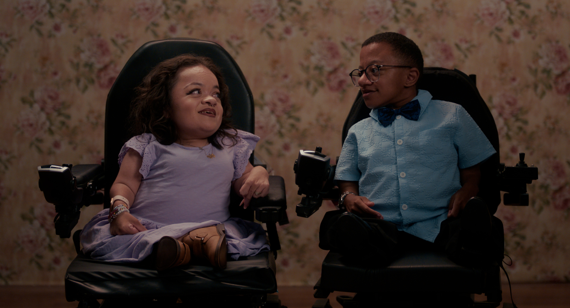 Pamela Chavez & Jay Manuel, two small people in power wheelchairs sit side by side against floral wallpaper, smiling at each other; Pamela wears a lavender dress and Jay a light blue shirt with a bow tie and glasses.